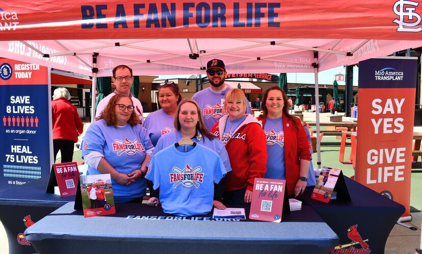 A group of seven Mid-America Transplant employees stand at a booth at Busch Stadium. They are wearing blue shirts that read "Fans for Life". Dan, a Triage Coordinator, stands in the back row wearing a ball cap and sunglasses.