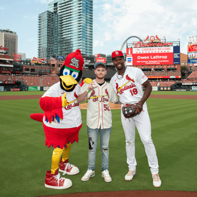 Owen Lathrop, wearing a Cardinals jersey, holds up a baseball at Busch Stadium. He is standing between Fredbird and player Jordan Walker.