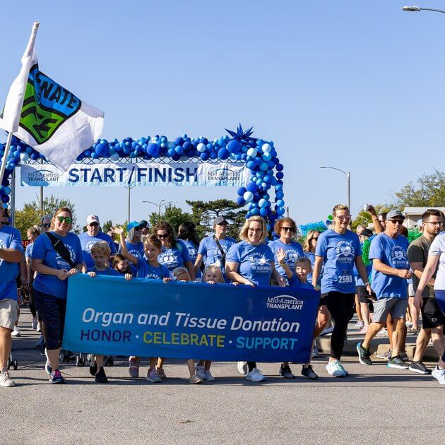 Donor families carry a banner that reads "Organ and tissue donation: Honor, Celebrate, Support". A start/finish sign is in the background and a Donate Life flag waves next to the walkers.
