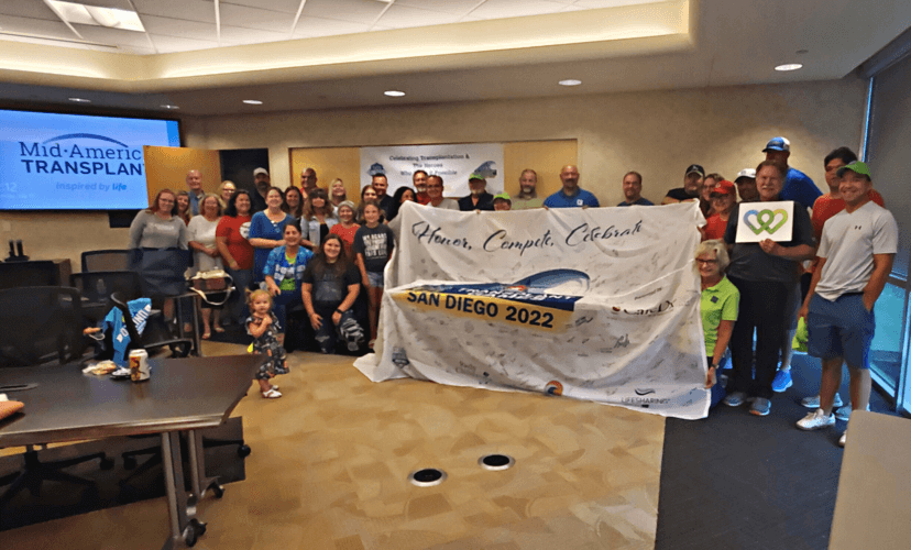 A group of people hold up a very large flag that reads "Honor. Compete. Celebrate. San Diego 2022"