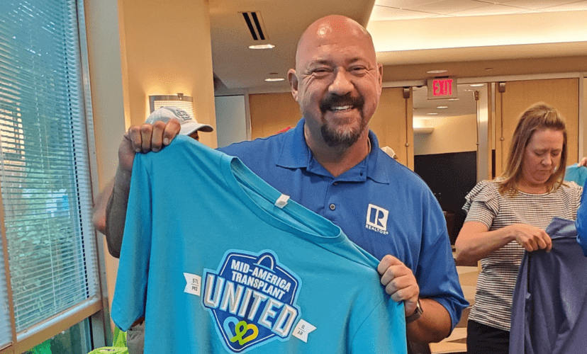 Will Klein smiles and holds up a blue shirt that reads "Mid-America Transplant United"