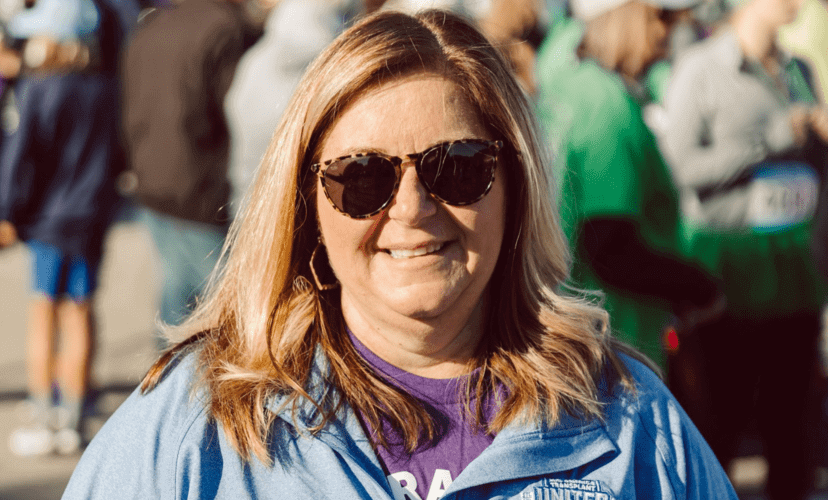 Lisa Taake wearing sunglasses, a purple shirt, and a blue jacket. She has blonde hair.