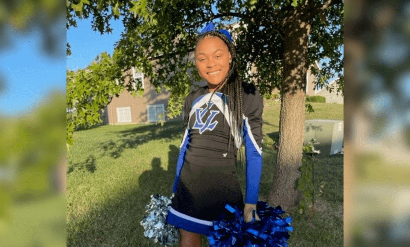 Jasmine Torrain wears a black and blue cheerleading uniform and pom-poms. She is posing in front of a tree.