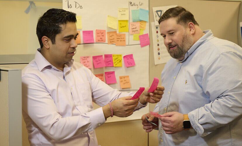 Vikram, a business intelligence employee, works with Louie from the Performance Excellence team to improve processes. Vikram is holding two pink post-it notes.