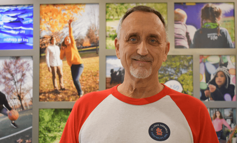 Keith, an employee at Mid-America Transplant, stands in the building's lobby. He is wearing a Fans for Life t-shirt and behind him is a photo collage of organ recipients.