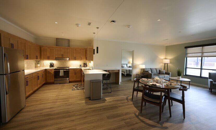 The interior of an apartment at the new Family House. The floor plan is open, with a kitchen on the left, a dining table in the center, and a comfortable living area on the right. A window on the far right brightens the room.