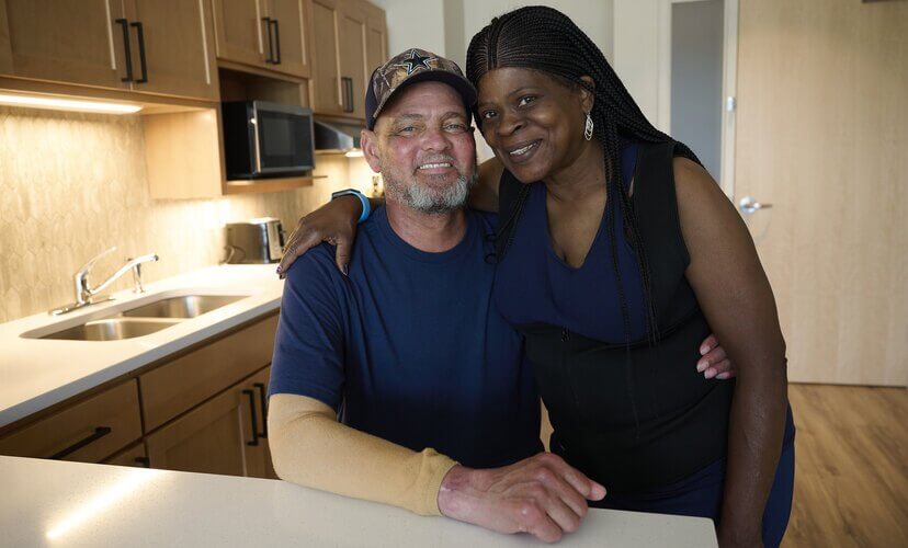 Lung recipient Bobby pictured with his wife Patricia. Bobby has a beard and wears a blue shirt with a camo hat. Patricia wears a blue shirt, a black vest, and has long beautiful braids.
