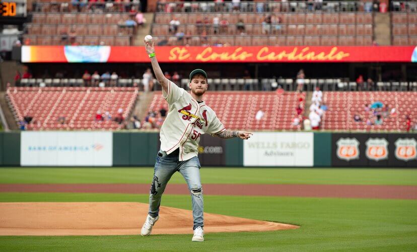 Owen Lathrop throws the first pitch at a Cardinals game. He is wearing a Cardinals jersey, jeans, and a green Donate Life bracelet.