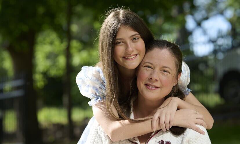 Lung recipient Paris hugs her mom Amy in the yard of the new Family House. Paris has long brown hair and wears a blue and white dress with puffy sleeves. Amy wears a white sweater.