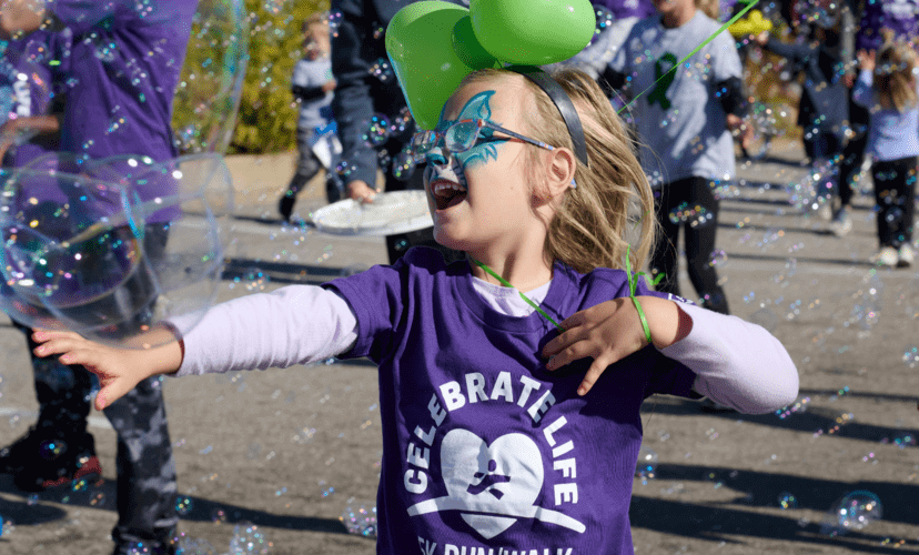 A young girl with blonde hair laughs into a stream of bubbles. Her face is painted with a blue butterfly.