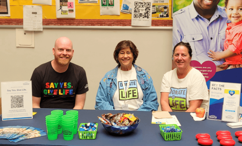 Dan is wearing a black Juneteenth shirt that says "Say Yes. Give Life." He is sitting at a table with two volunteers wearing Donate Life shirts. The table has Mid-America Transplant promotional items on it.