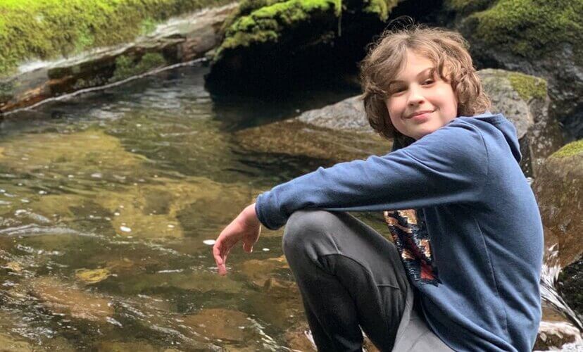 Beckham, a teenager with brown hair and a blue hoodie, sits on a rock next to a creek