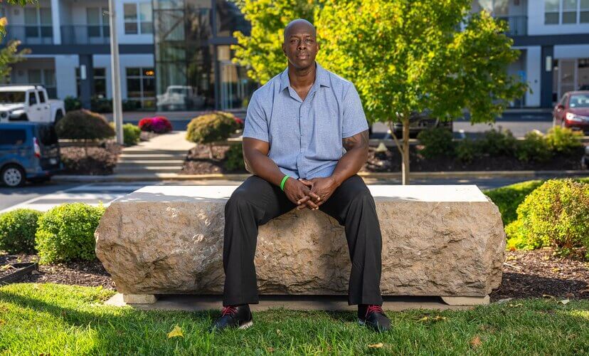 A man sits on a stone bench with buildings and trees in the background. It's a sunny day. He is wearing a blue collared shirt and a green Donate Life bracelet.