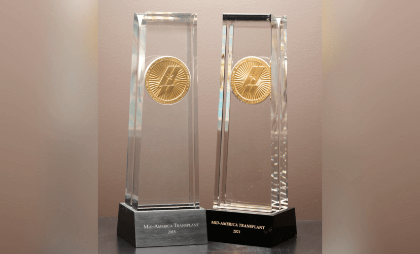 Two awards featuring the Malcolm Baldrige logo stand together. Both read "Mid-America Transplant" on the base. The left one has a grey base and is marked 2015. The right one has a black base and is labeled 2021.