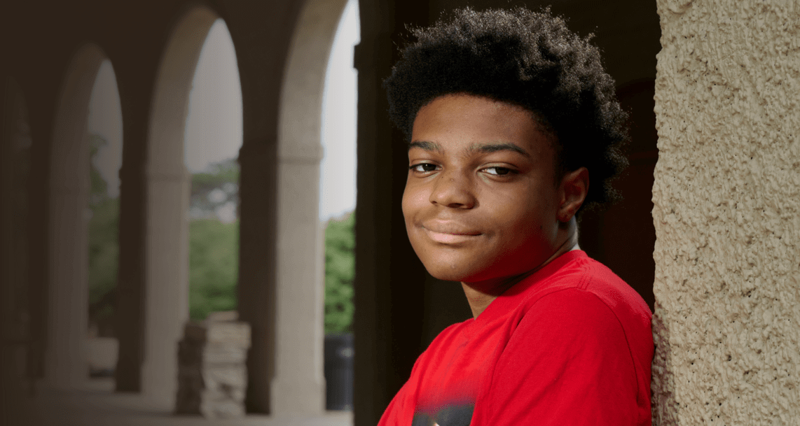 A teenage boy in a red shirt leans against a tan wall