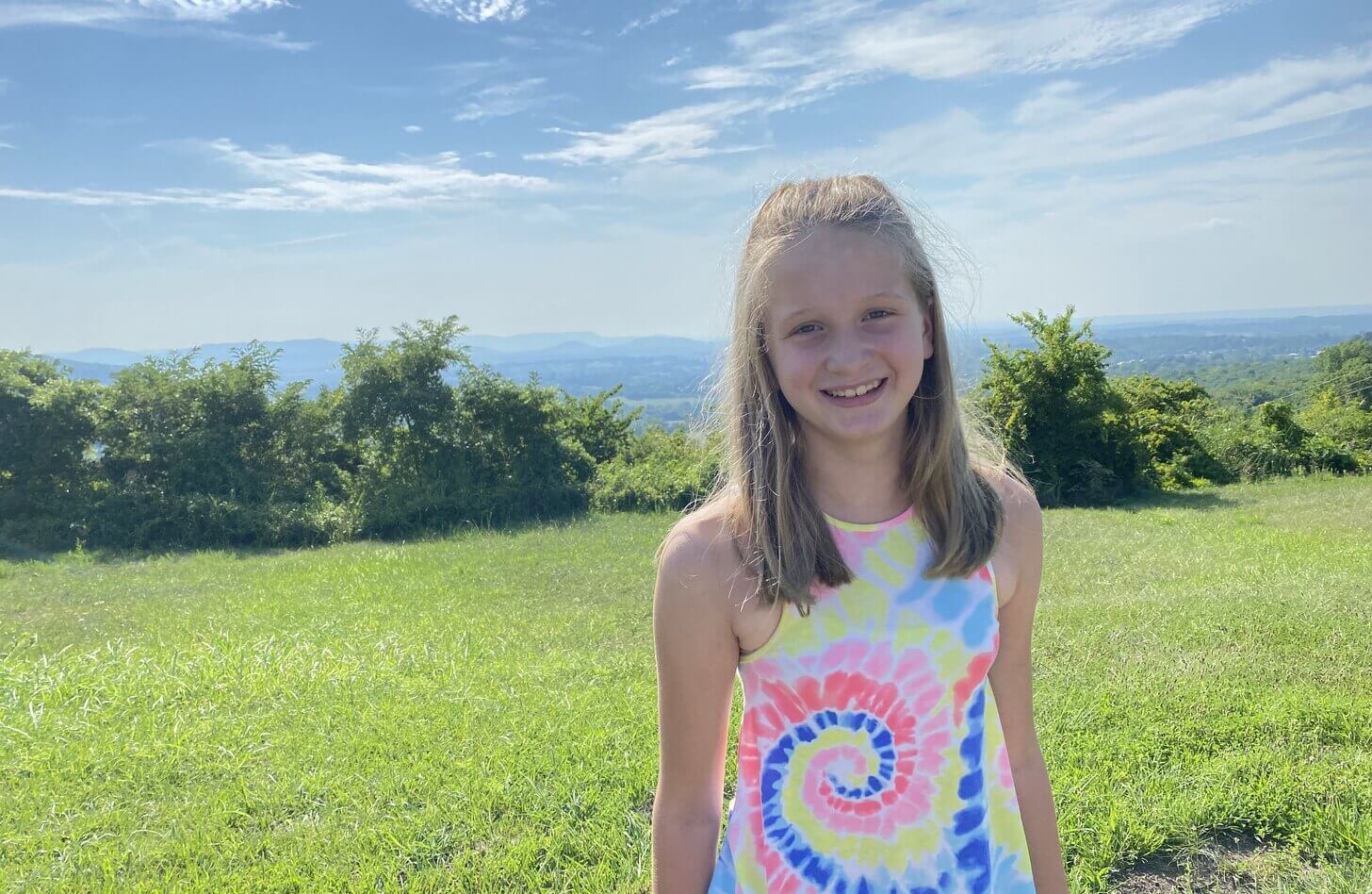 A girl in a tye-dye shirt stands in front of a green field and a blue sky