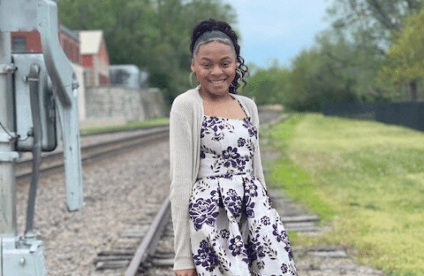 A teenage girl wearing a white dress with purple flowers on it and a beige cardigan. She is posing next to train tracks.