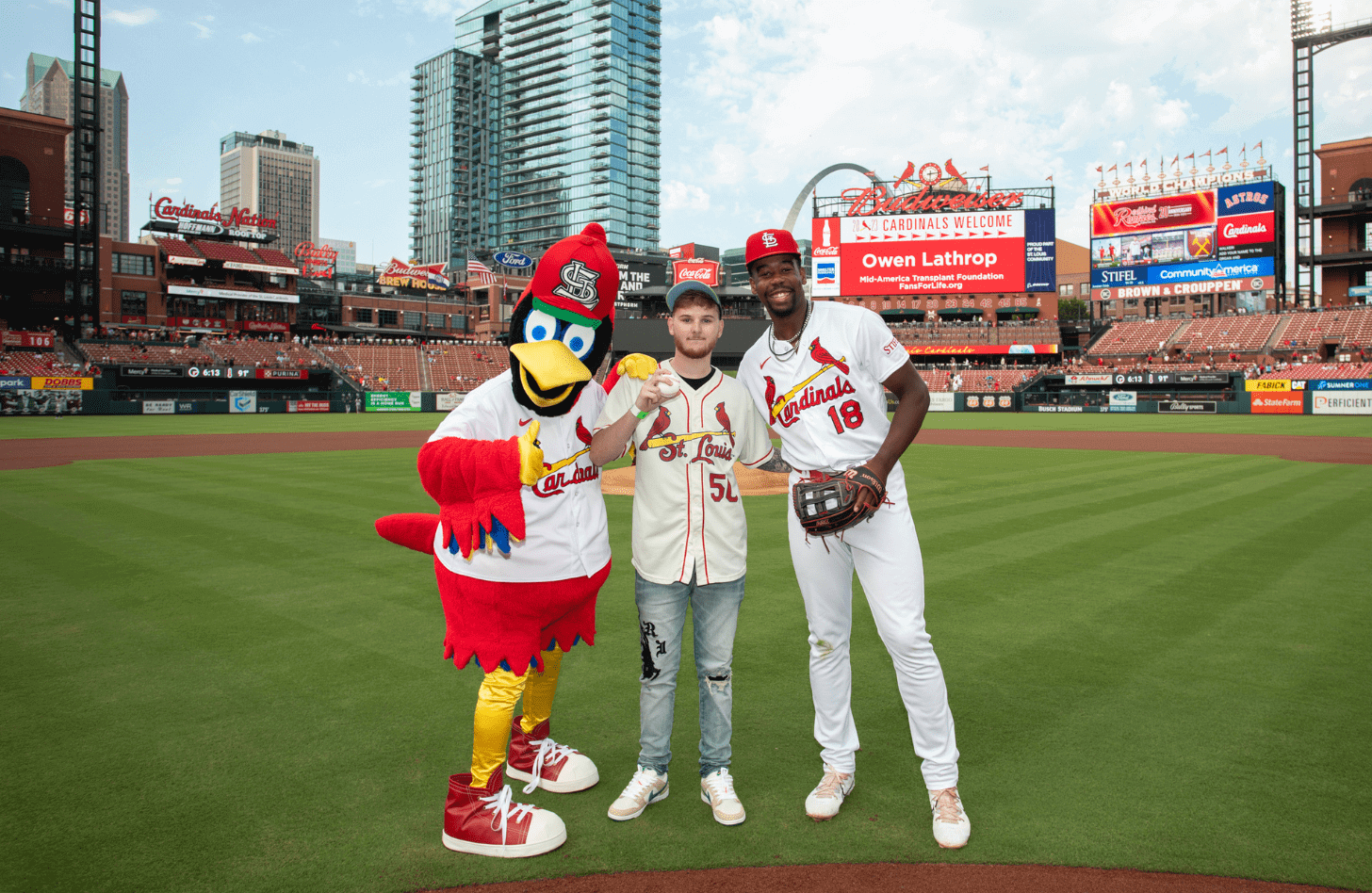 Owen Lathrop, wearing a Cardinals jersey, holds up a baseball at Busch Stadium. He is standing between Fredbird and player Jordan Walker.