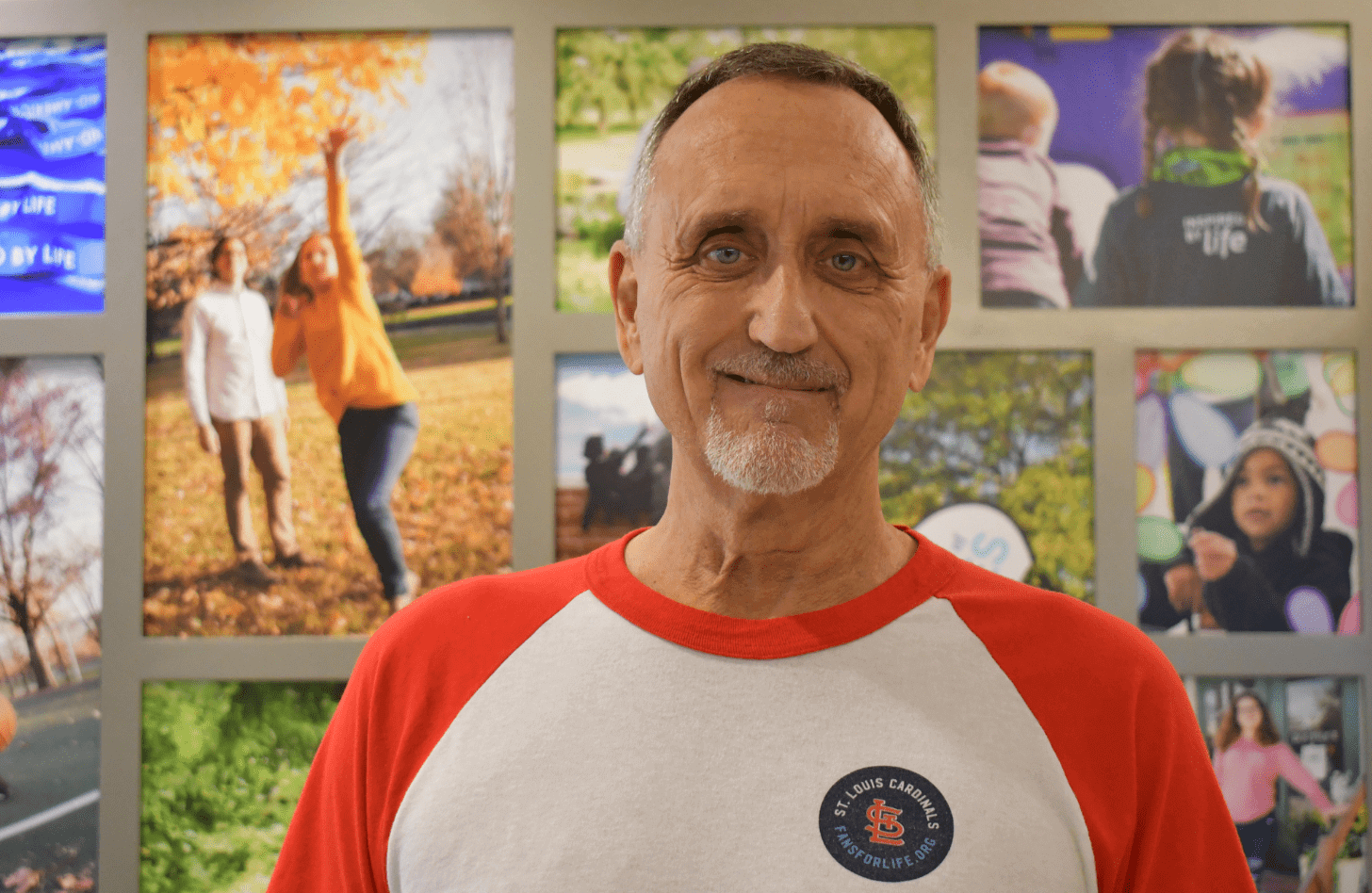 Keith, an employee at Mid-America Transplant, stands in the building's lobby. He is wearing a Fans for Life t-shirt and behind him is a photo collage of organ recipients.