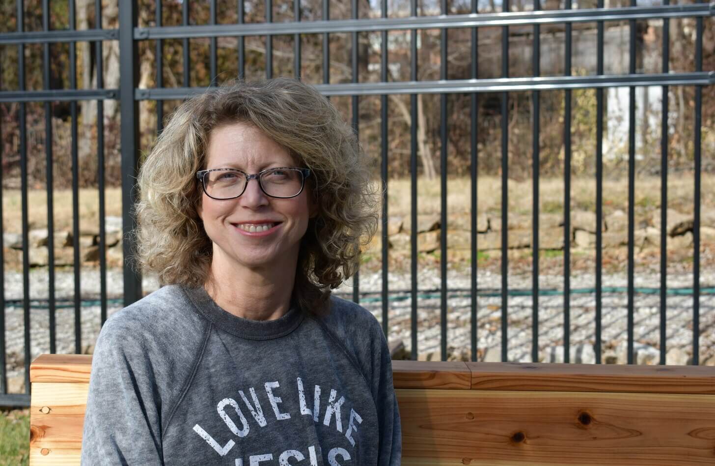 A woman with wavy greying blonde hair sits on a bench. Her grey sweatshirt reads "love like Jesus"