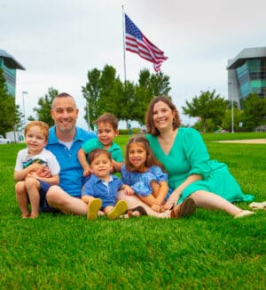 A family sits in the grass. A man, a woman, and four young kids. An American flag waves in the background.