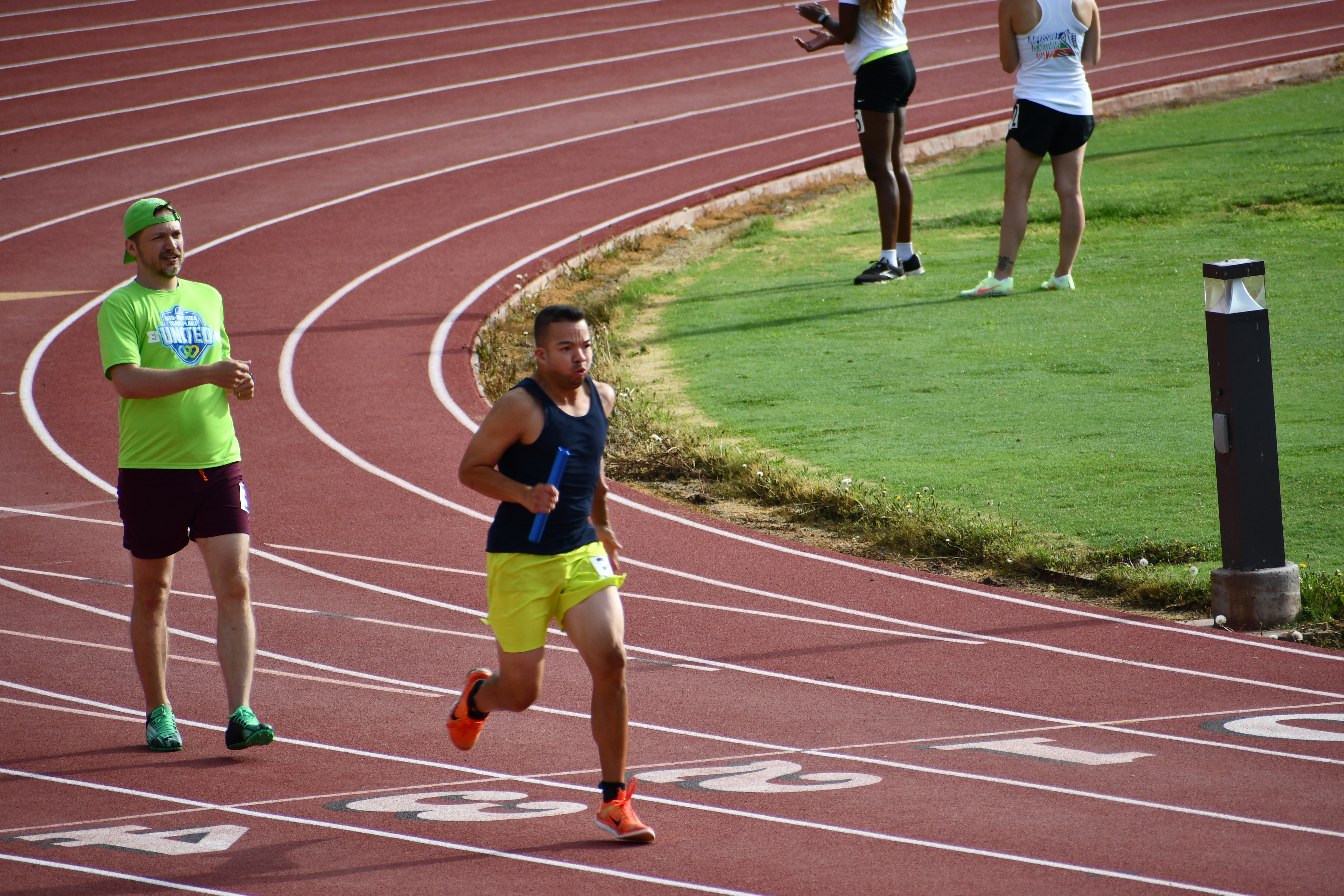 Joe, wearing neon yellow shorts and a navy tank top, running a relay race at the 2022 Transplant Games of America. His teammate behind him is wearing an MT United shirt.