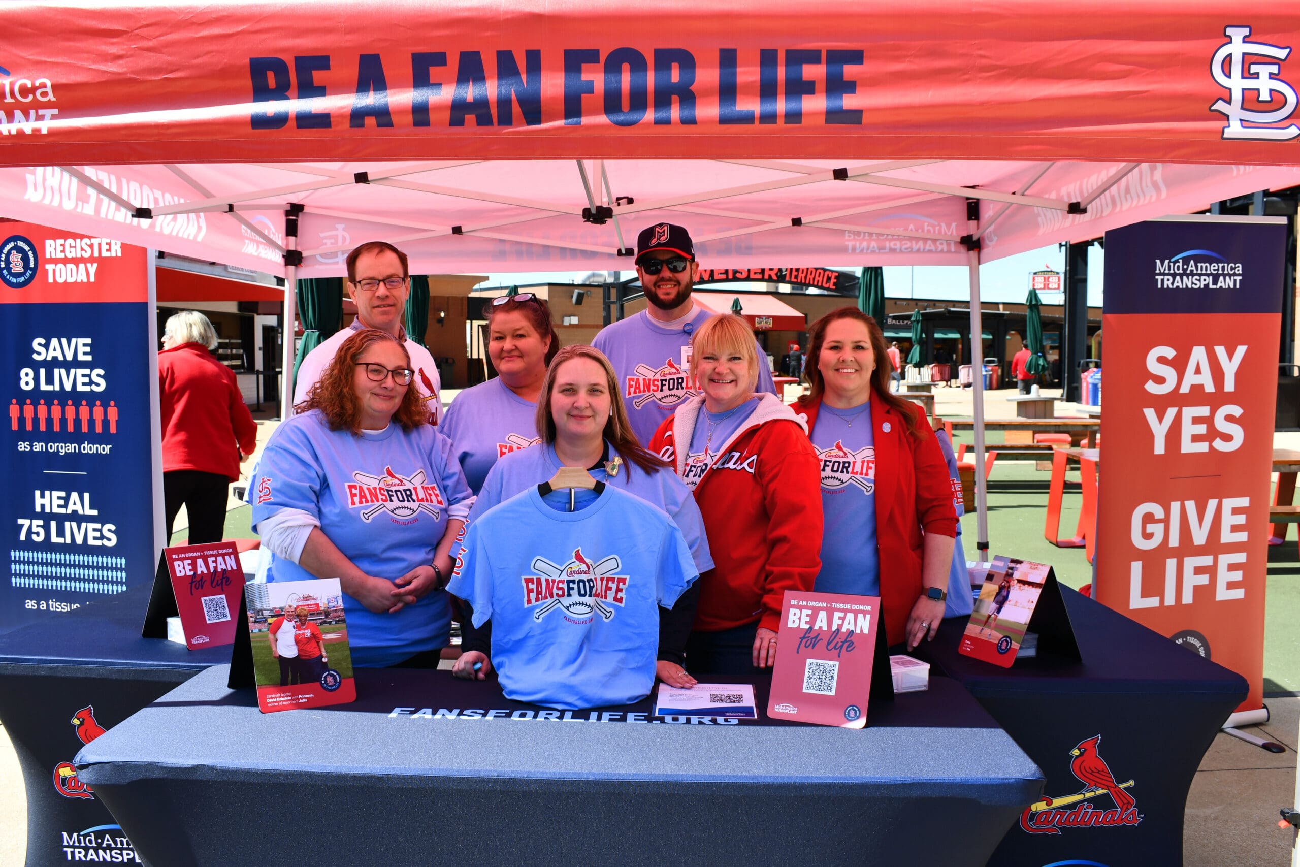 A group of seven Mid-America Transplant employees stand at a booth at Busch Stadium. They are wearing blue shirts that read "Fans for Life". Dan, a Triage Coordinator, stands in the back row wearing a ball cap and sunglasses.