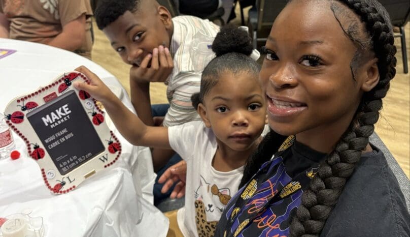 A family sits at a table to work on a memory-making craft project. A little girl holds up a photo frame while her brother leans on the table behind her, his chin in his hand.